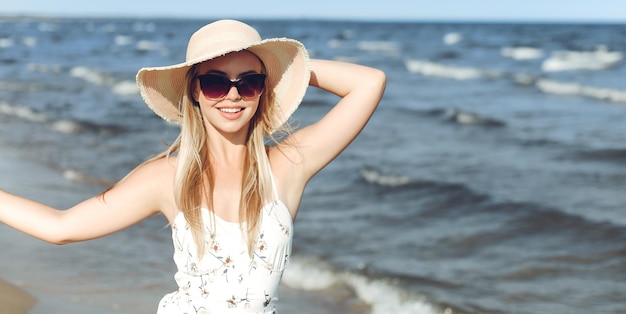 Glückliche blonde Frau in freier Glückseligkeit am Meerstrand mit Sonnenbrille und Hut.