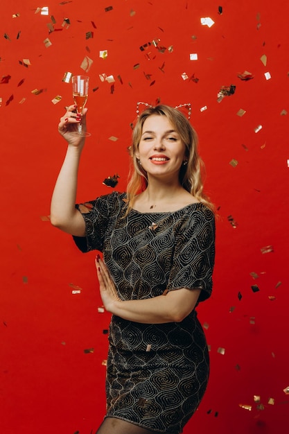 Glückliche blonde Frau in glänzender Modekleidung ist glücklich, ein Glas Champagner zu halten und mit Konfetti auf rotem Hintergrund zu posieren