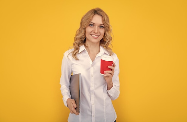 Glückliche blonde Frau mit Kaffeetasse und Laptop auf gelbem Hintergrund