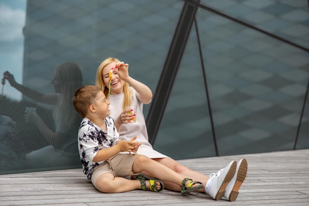 Glückliche blonde Frau und kleiner Junge, die auf der Terrasse sitzen und Süßigkeiten essen Mutter und Sohn genießen die gemeinsame Zeit Positive junge Mutter spielt und verbringt Zeit mit ihrem süßen Kind, das lacht und Spaß hat Familie