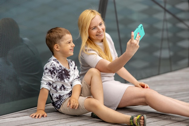 Glückliche blonde Frau und kleiner Junge sitzen auf der Terrasse und machen Selfie auf dem Smartphone Mutter und Sohn genießen die gemeinsame Zeit Positive junge Mutter verbringt Zeit mit ihrem süßen Kind Spaß haben Familie