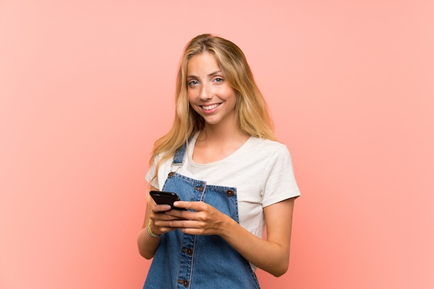 Glückliche blonde junge Frau mit einem Handy über lokalisierter rosa Wand