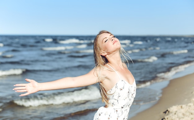 Glückliche blonde schöne Frau am Meeresstrand, die in einem weißen Sommerkleid steht und die Hände hebt.