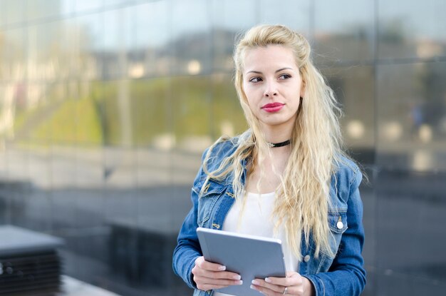 Glückliche blonde Studentin mit Tablette in einer reflektierenden Wand