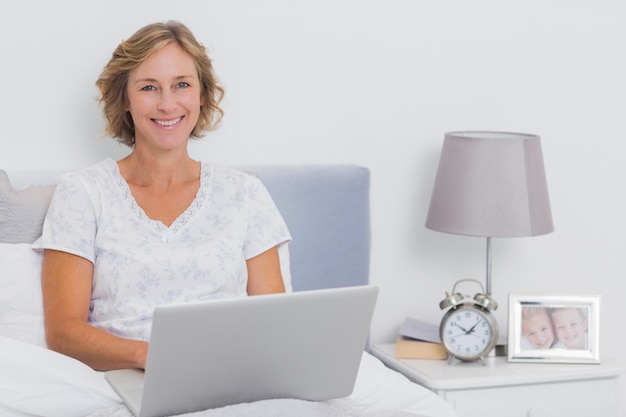 Glückliche Blondine, die im Bett unter Verwendung des Laptops sitzen