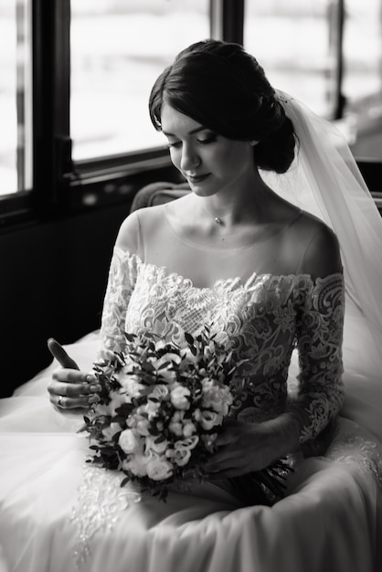 Glückliche Braut, die nahe Fenster sitzt und den Hochzeitsstrauß betrachtet. Schöne junge Frau in einem weißen Kleid mit einem Blumenstrauß.