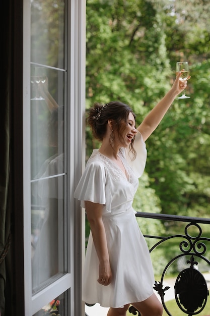 Glückliche Braut junge Frau in einem seidenen kurzen Peignoir wirft ein Glas Champagner lächelt und schaut auf den B...