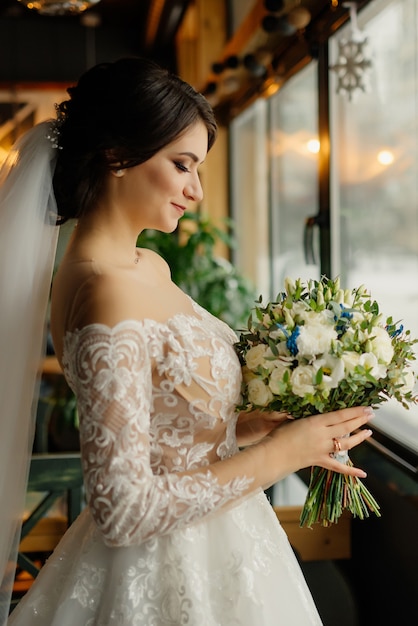 Glückliche Braut steht im Dachbodeninneren nahe Fenster und schaut auf den Hochzeitsstrauß. Schöne junge Frau in einem weißen Kleid mit einem Blumenstrauß.