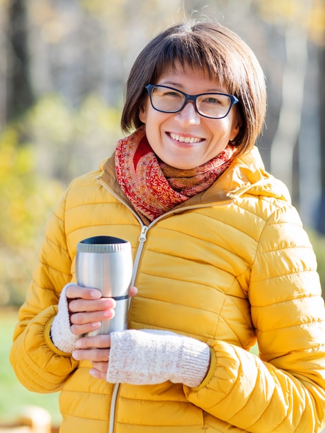 Glückliche breite lächelnde Frauen im hellen gelben jacketis, der Thermosbecher hält. Heißer Tee am kühlen Herbsttag.