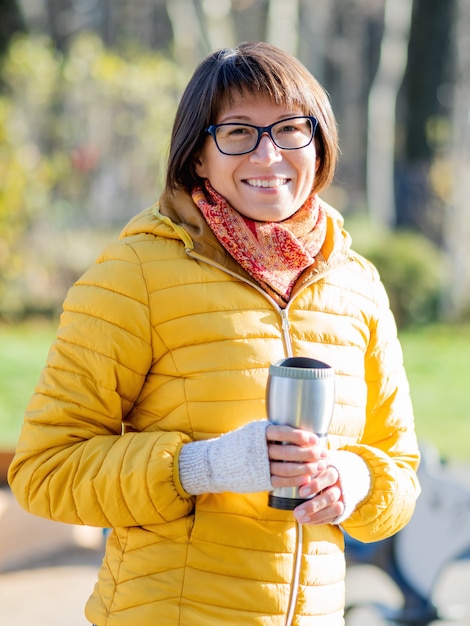 Glückliche breite lächelnde Frauen im hellen gelben jacketis, der Thermosbecher hält