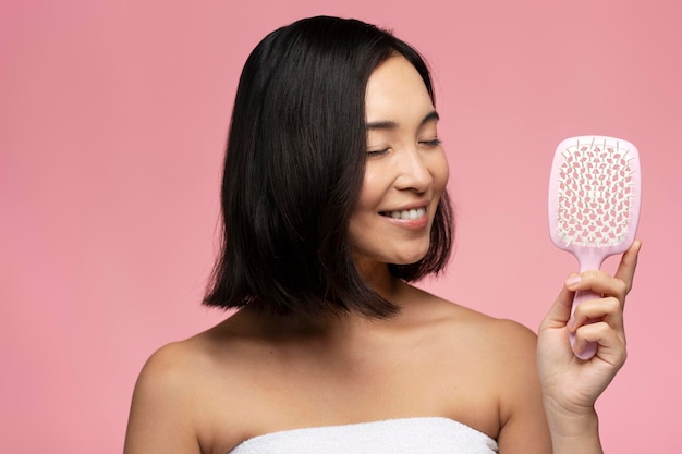 Glückliche brünette Asiatin mit seidigem Haar, die moderne Haarbürste isoliert hält
