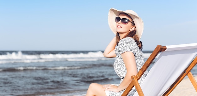 Glückliche brünette Frau mit Sonnenbrille beim Entspannen auf einem hölzernen Liegestuhl am Meeresstrand