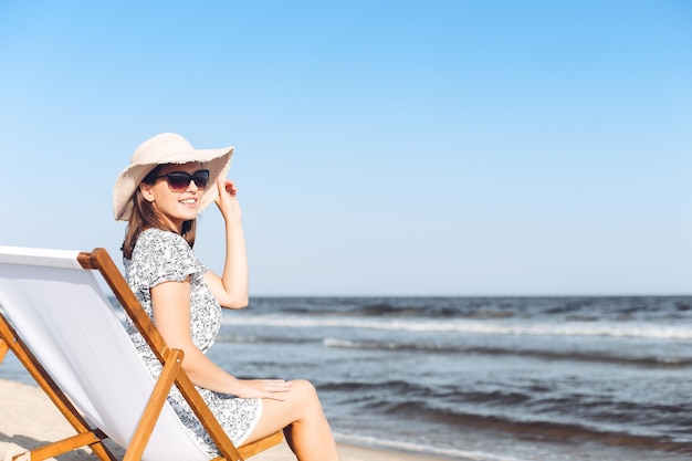 Glückliche brünette Frau mit Sonnenbrille beim Entspannen auf einem hölzernen Liegestuhl am Meeresstrand