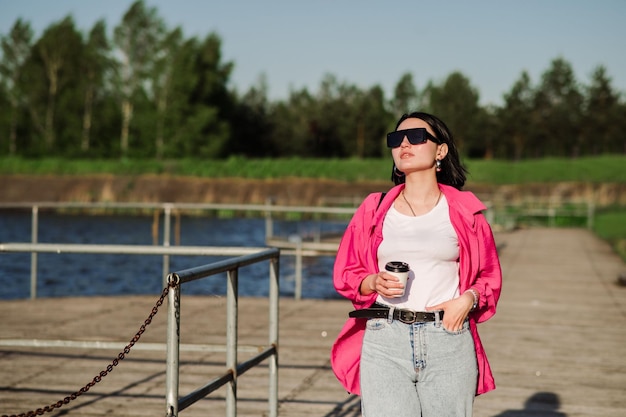 Glückliche brünette Frau mit Sonnenbrille, die an sonnigen Tagen in der Nähe des Sees auf einem Holzsteg spazieren geht