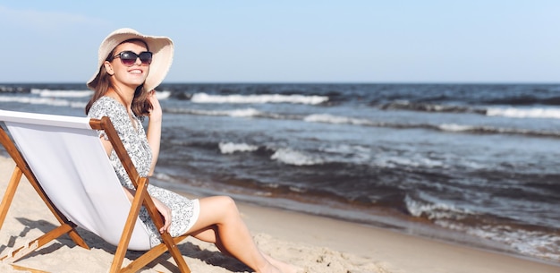 Glückliche brünette Frau mit Sonnenbrille und Hut entspannt sich auf einem hölzernen Liegestuhl am Meeresstrand