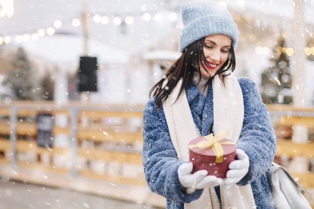 Glückliche brünette Frau trägt Strickmütze und blauen Mantel mit roter Geschenkbox auf dem Weihnachtsmarkt während des Schneefalls. Platz für Text