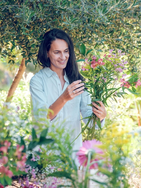 Glückliche Brünette in weißem Hemd, die an einem sonnigen Tag in der Natur einen Blumenstrauß aus sanften Blumen herstellt