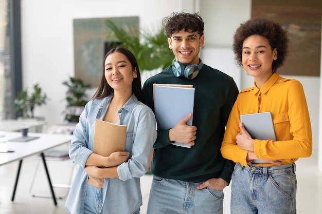 Glückliche drei gemischtrassige Studenten posieren im Coworking Space, junge Leute mit darin stehenden Arbeitsbüchern