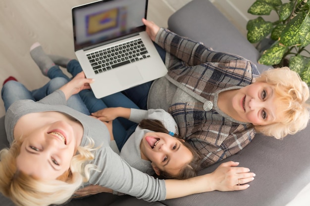 Glückliche drei Generationen von Frauen sitzen entspannt auf der Couch und lachen lustige Videos auf dem Laptop an, lächeln positive Frauen, Großmutter, Mutter und Tochter haben Spaß, ruhen sich auf dem Sofa aus und genießen den Film auf dem Computer