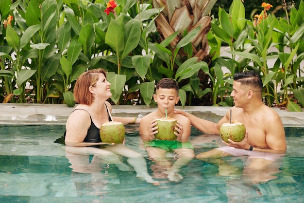 Glückliche dreiköpfige Familie, die im Schwimmbad sitzt und erfrischende Kokosnusscocktails trinkt