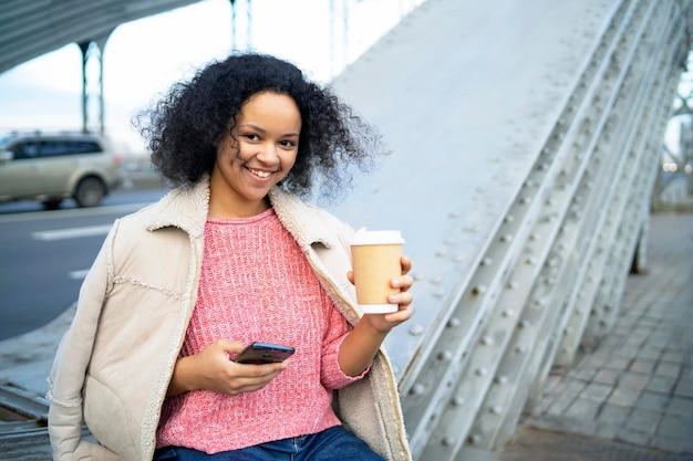 Glückliche dunkelhäutige frau, die ein mobiles smartphone in ihren händen hält afroamerikanische frau, die kaffee trinkt...