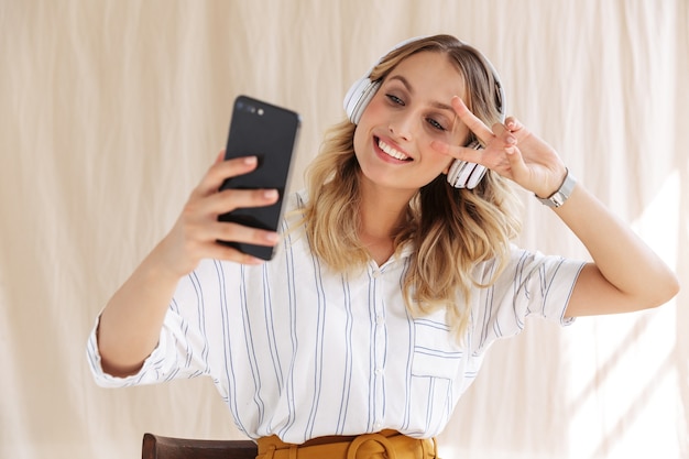 glückliche, elegante blonde Frau mit Kopfhörern, die ein Selfie-Foto auf dem Smartphone macht