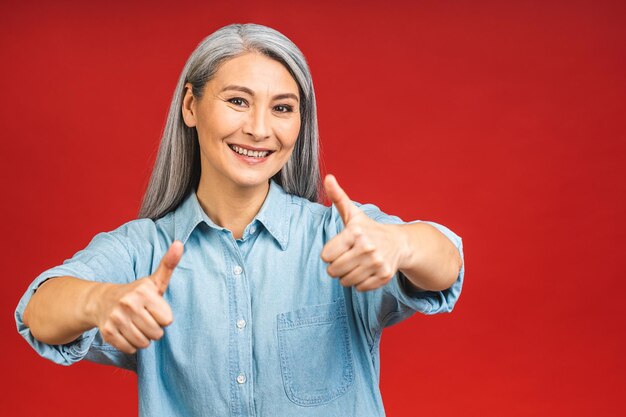 Glückliche, elegante, reife Senioren im Alter von asiatischen Geschäftsfrauen, die über rotem Hintergrund isoliert stehen Lächelnde, selbstbewusste, fröhliche Dame mittleren Alters, Kopienraum Daumen hoch Zeichen