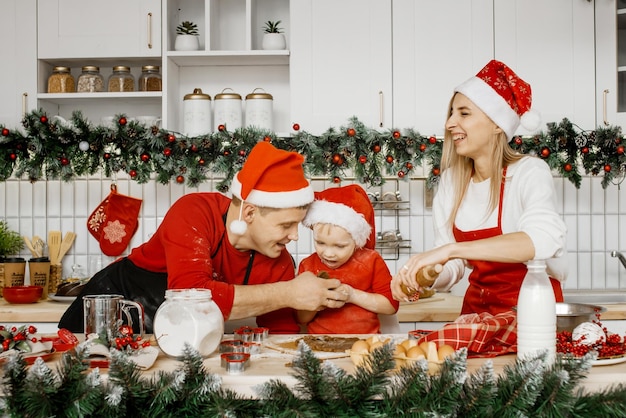 Glückliche Eltern in Weihnachtsmützen und kleiner Sohn sind in der Küche und bereiten Teig zum Backen von Weihnachtsplätzchen vor. Spaß haben, eine glückliche Familie zu spielen, die auf die Feiertage wartet