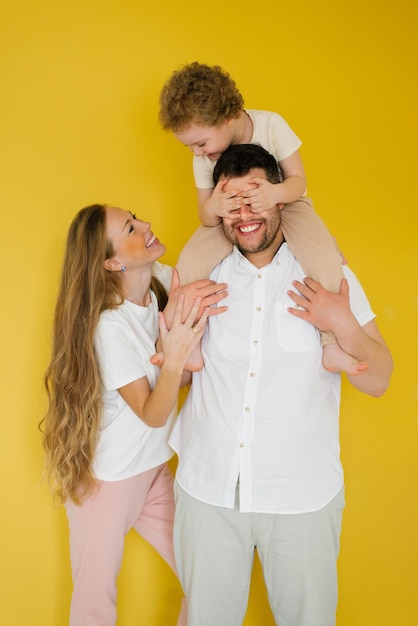 Glückliche Eltern umarmen ihren Sohn, der Liebe warme Gefühle ausdrückt Das Kind sitzt auf dem Hals seines Vaters und bedeckt seine Augen mit seinen Handflächen Spaß Familie isoliert gelber Hintergrund