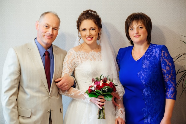 Glückliche Eltern und die Tochter, die heiratet, gekleidet in ein Hochzeitskleid und einen Blumenstrauß in der Hand