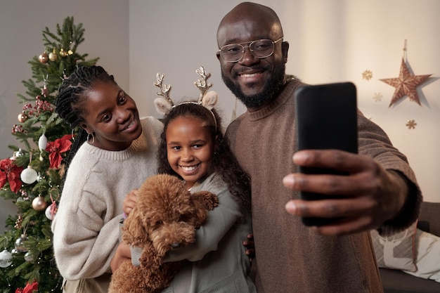 Glückliche Eltern und ihre süße Tochter mit Haustier, die in die Smartphone-Kamera schaut, während sie am Weihnachtstag ein Selfie macht