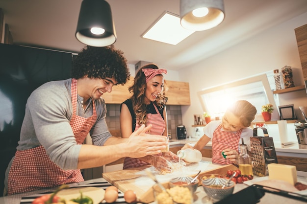 Glückliche eltern und ihre tochter bereiten gemeinsam in der küche teig für pizza zu. Kleines Mädchen hilft ihren Eltern, Milch in das Mehlbündel auf dem Tisch zu gießen.