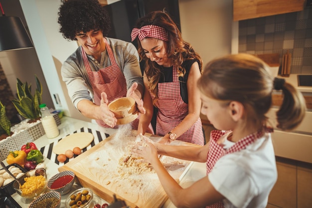 Glückliche eltern und ihre tochter bereiten gemeinsam in der küche teig für pizza zu. Kleines Mädchen hilft ihren Eltern, Teig auf dem Tisch zu mischen.