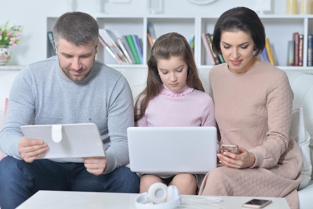 Glückliche Eltern und Tochter, die digitale Geräte benutzen, während sie auf dem Sofa im Zimmer sitzen