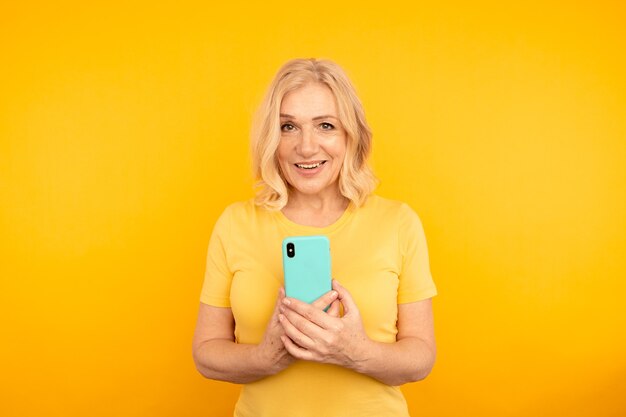 Glückliche erwachsene Frau mit blauem Handy auf dem gelben Hintergrund.