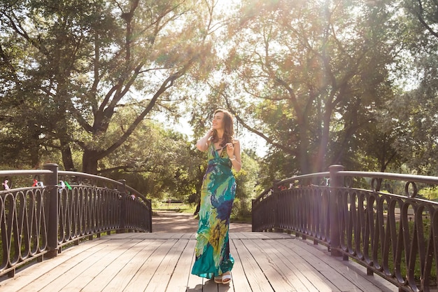 Glückliche erwachsene Frau mittleren Alters, die den Sommer im Park genießt, geht über die Brücke im Freien, gekleidet in grünes, modisches Kleid, das wegschaut