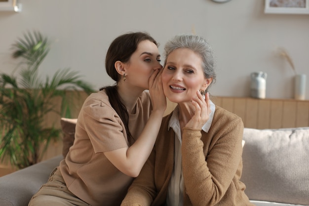 Glückliche erwachsene Tochter flüstert ihrer mittleren Mutter zu Hause Geheimnis zu, klatscht, teilt Geheimnis. Schöne Frau, die gute Nachrichten von reifer Mutter, vertrauenswürdigen Beziehungen hört.