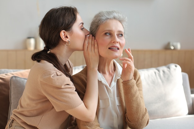 Glückliche erwachsene Tochter flüstert ihrer mittleren Mutter zu Hause Geheimnis zu, klatscht, teilt Geheimnis. Schöne Frau, die gute Nachrichten von reifer Mutter, vertrauenswürdigen Beziehungen hört.