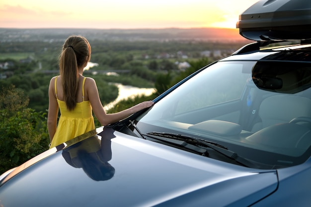 Glückliche Fahrerin im Sommerkleid, die einen warmen Abend in der Nähe ihres Autos genießt. Reise- und Urlaubskonzept.