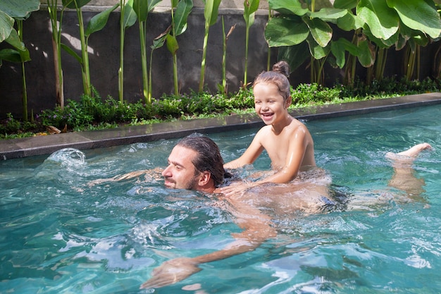 Glückliche Familie, aktiver Vater mit kleinem Kind, entzückendes Kleinkindmädchen, Spaß zusammen im Außenpool im Wasserpark während des sonnigen Sommerurlaubs im tropischen Resort.