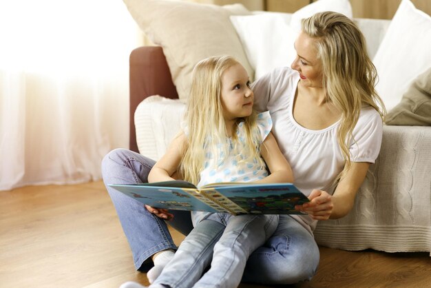 Glückliche Familie. Blonde junge Mutter liest ihrer süßen Tochter ein Buch vor, während sie auf dem Holzboden in einem sonnigen Zimmer sitzt. Mutterschaftskonzept.