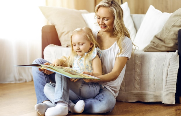 Glückliche Familie. Blonde junge Mutter liest ihrer süßen Tochter ein Buch vor, während sie im sonnigen Zimmer auf Holzboden sitzt. Mutterschaftskonzept.