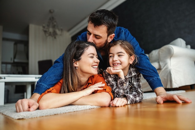 Glückliche Familie, die eine tolle Zeit zusammen hat. Mama und Tochter liegen auf dem Boden, während Papa Mama küsst. gesunde Kindheit.