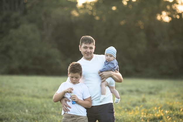 Glückliche Familie, die sich auf einem Sommerspaziergang umarmt und umarmt. Vater und seine beiden Söhne gehen im Park spazieren und genießen die wunderschöne Natur. Niedliches Familienporträt