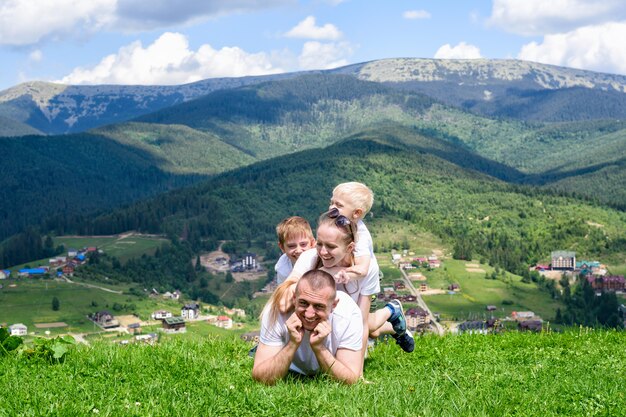 Glückliche Familie: froher Vater, Mutter und zwei Söhne liegen auf dem grünen Gras der Wald, die Berge und der Himmel mit Wolken.