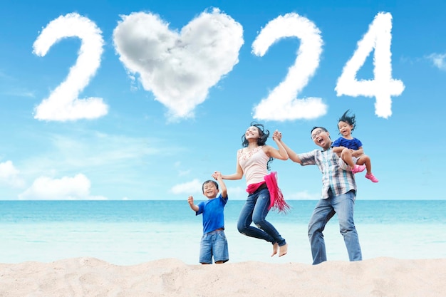 Glückliche Familie genießt den Urlaub am Strand mit wolkenförmigen Neujahrszahlen 2024 und einem Herzen am Himmel
