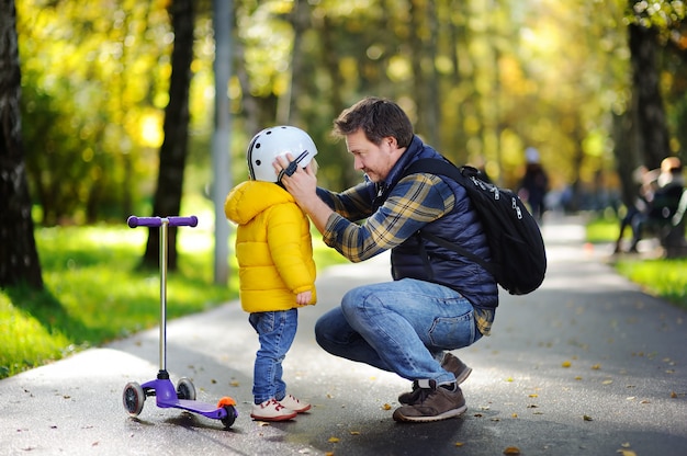 Glückliche familie im herbstpark. mittelaltervater, der seinem kleinen sohn hilft, seinen sturzhelm zu setzen. aktiver kleinkindjunge, zum eines rollers zu reiten. kindersicherheit