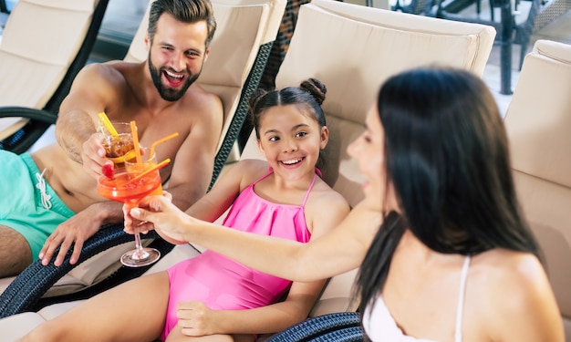 Glückliche Familie im Resort. Der hübsche Vater, die schöne Mutter und die kleine süße Tochter liegen auf den Sonnenliegen im großen Wellnesscenter mit Pool und trinken Saft und Cocktails