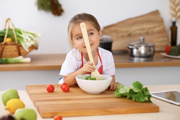 Glückliche Familie in der Küche Mutter und Tochter kochen leckeres Frühstück mit frischem Salat Kleiner Helfer, der Tomaten und Grün schneidet und mischt