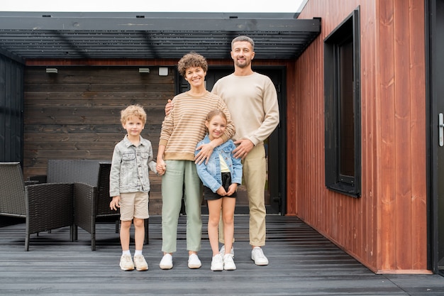 Glückliche Familie junger Eltern und zwei kleine Kinder, die auf dem Holzboden der Terrasse ihres neuen Hauses mit schwarzen Sesseln dahinter stehen